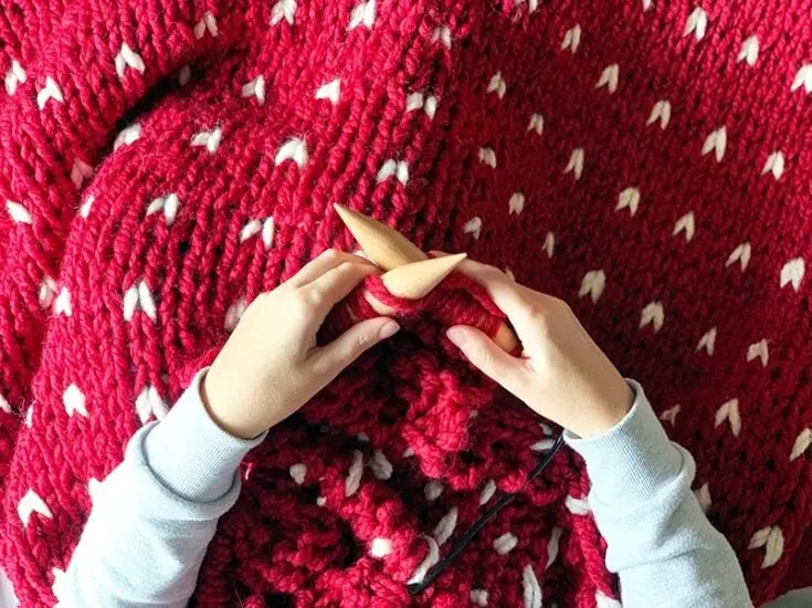 woman's hands and forearm knitting red and white patterned blanket with large knitting needles